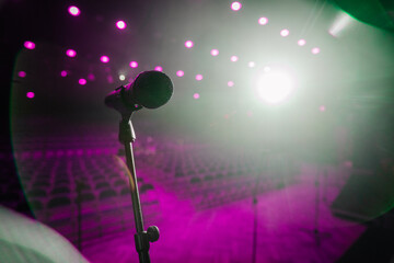 A microphone on a stand in a spotlight on an empty stage, with blurred audience seating in the background, creating a dramatic atmosphere..