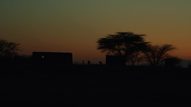 Kenya, Loima - September 9, 2023: Boy plays and jumps around man sitting in chair. View of houses in settlement in savannah and beautiful landscape with mountains silhouetted against evening sky