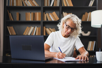 Middle aged senior woman using laptop computer writing notes at workplace. Focused mature old...