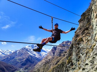 With arms wide open, an individual enjoys the exhilaration of crossing a rope bridge amidst snow-covered mountains under a clear blue sky.