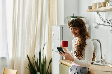 A young woman smiles as she holds a red cup and checks her phone in a bright kitchen space
