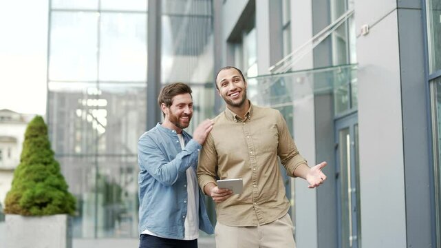 Two happy business colleagues have friendly conversation walking outside office building. Joyful worker praises friend for good results, achievements, congratulates shaking hands, smiling, rejoicing