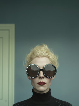 Elegant woman with platinum blonde hair wearing round dark sunglasses and a black turtleneck, standing in front of a muted blue wall.