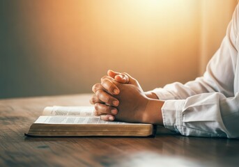 Praying hands resting on open bible, warm sunlight illuminating wooden surface, expressing spiritual reflection and deep religious connection
