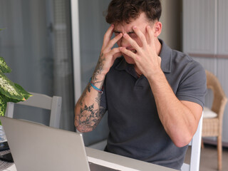 Young angry guy with hands on face while working on laptop expressing annoyance, dissatisfaction....