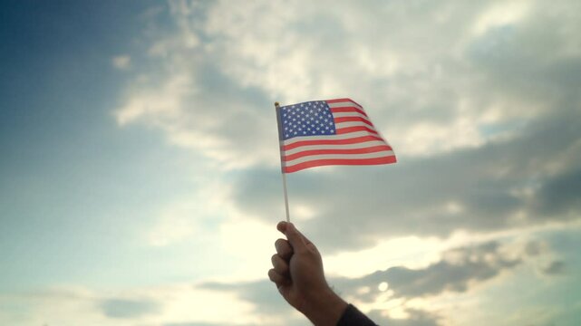 Close up unrecognized man's holding USA flag on one hand against sky with sunset light background, proud american holding the United states flag celebration.
