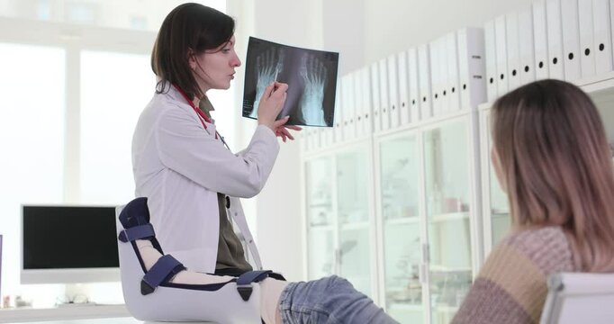 Female doctor shows X-ray images of feet to lady patient with leg in cast. Traumatologist controls injury recovery of young woman in hospital office