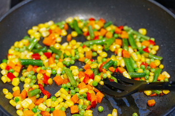 A colorful medley of corn, peas, carrots, green beans, and red peppers sizzles in a black pan, stirred with a black spatula, showcasing a simple, healthy side dish.