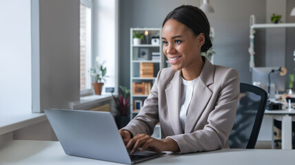 A happy young African American businesswoman working on a laptop in a modern office with a smile, wearing a professional beige blazer and white blouse