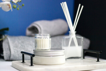 A cream, candle, and diffuser on a marble tray against a blue background, accompanied by soft towels, perfect for spa treatments, body care, and relaxation.