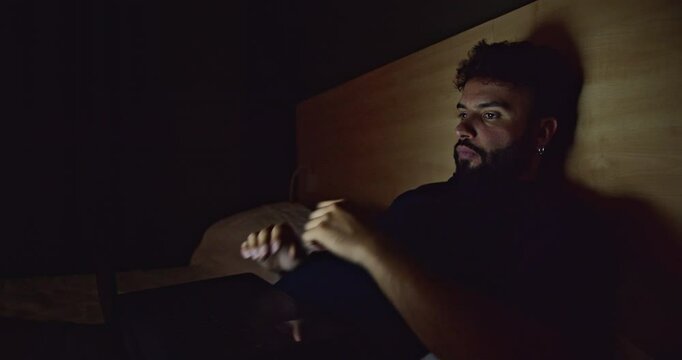 A young Caucasian man lies in bed at night, looking at his laptop. He is illuminated by the light of the screen.
