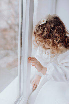 cute little girl with curls sitting on windowsill, looking out window. safety of small children in apartment, precautions against falling for toddlers on high floors, no face