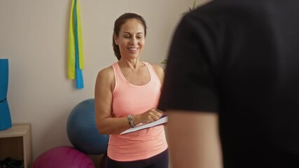 Middle-aged hispanic woman in gym, wearing pink tank, smiling and holding clipboard, extends hand for handshake, in indoor sports center with exercise equipment, talking to unseen man.