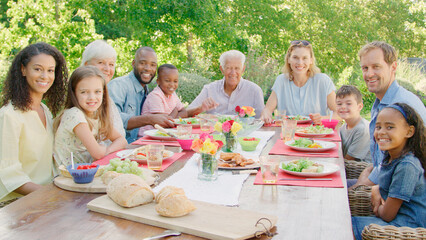 Portrait Of Multi-Generation Family With Friends Sitting Around Outdoor Table In Garden Enjoying Meal Together