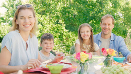 Portrait Of Family Sitting Around Outdoor Table In Summer Garden Enjoying Meal Together