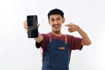 Young handsome asian barista man wearing apron pointing finger at mobile screen, showing app and smiling, standing over white background