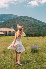 Young Blonde haired Woman in white dress walking on the green grass of Southern France, in front of Mountains