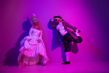 Elegant couple in 18th-century costumes posing under vibrant neon lighting during a themed masquerade ball or carnival night.
