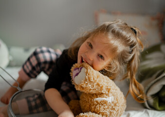 Happy little girl playing and chewing her teddy bear in the morning wearing pajamas. Child girl...