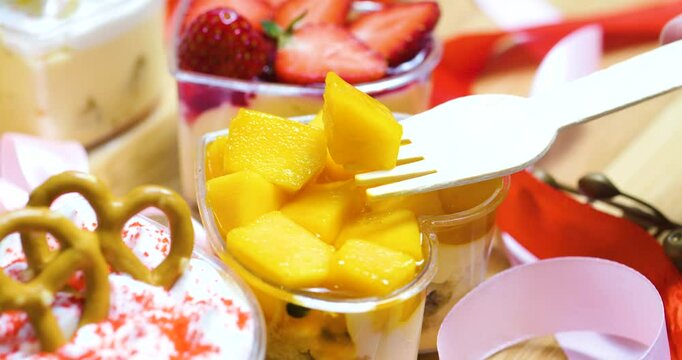 Wooden fork lifting juicy mango chunks from heart shaped container, surrounded by colorful desserts and decorative ribbons, highlighting tropical treat's elegance