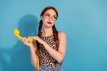 Stylish young woman with red braids wearing a leopard print top and glasses holding a yellow phone handset against a blue background