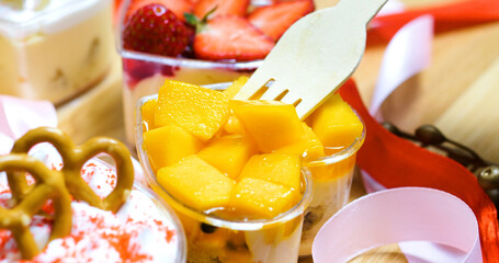 Wooden utensil lifting vibrant mango piece from heart shaped dessert, nestled amid elegant sweets, ribbons, festive decorative elements