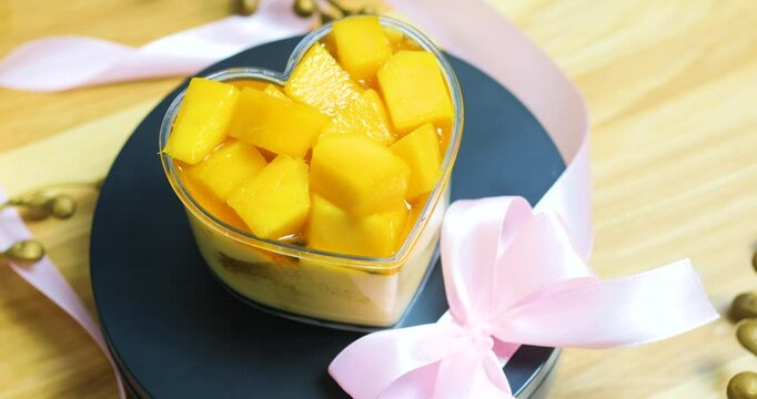 Heart shaped mango mousse garnished with fresh mango pieces, adorned by soft pink ribbon, sitting atop dark ceramic plate against rustic wooden surface