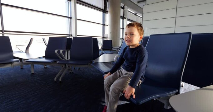 Young toddler waiting at airport gate, sitting in chair and moving legs energetically, while growing increasingly restless. Little boy speak and look around empty hall, playful and impatient