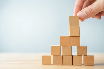 Obraz premium A hand places a wooden block atop a pyramid of smaller blocks on a light-colored surface, showcasing creativity and construction, Best Job Candidate.
