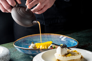 A chef elegantly pours a flavorful sauce over a beautifully presented dessert featuring fresh fruits and cream, showcasing a modern culinary art form.