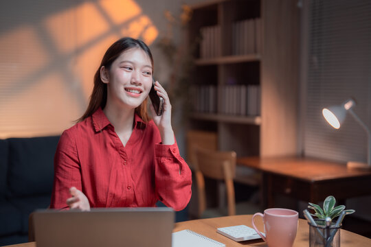 Asian female entrepreneur managing her business from home, working late at night on a laptop while talking on the phone, sitting at her desk in a cozy and well-lit environment
