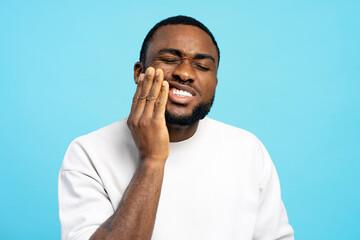Young man suffering from strong toothache touching his cheek on blue background