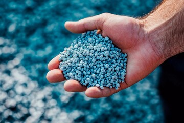 A hand is filled with small blue granules under sunlight, indicating a material used in industrial or agricultural applications. The background features blurred textures of similar granules