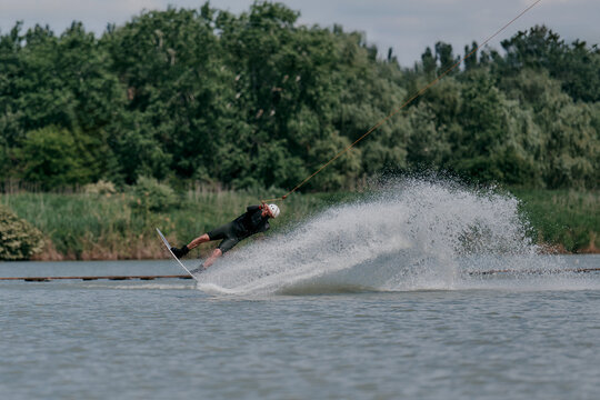 A man on a wakeboard on the lake does a backroll trick, creates splashes of water. Greenery and cloudy sky in the background