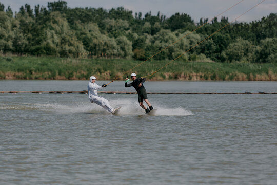 Two people are wakeboarding on a lake with a cable car system, walking next to each other in helmets and life jackets, surrounded by greenery and a cloudy sky
