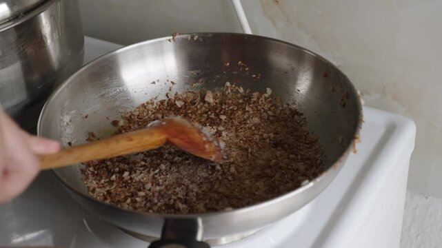 Person rubs wooden spatula over food burnt to steel frying pan on electric stove. Problems with cooking due to lack of non stick coating. Lifestyle and life difficulties in everyday life.