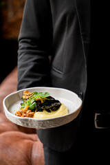 A person in a black outfit holds a beautifully plated dish featuring creamy mashed potatoes and vibrant vegetables, highlighting an elegant dining experience.