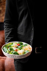 A close-up of a person holding a bowl of fresh salad with greens, cherry tomatoes, and colorful shredded vegetables, set against a stylish backdrop.