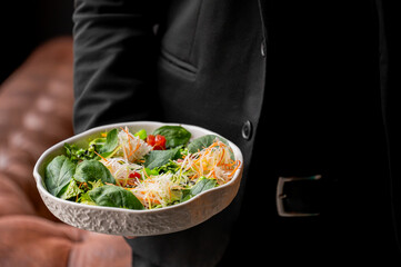 A close-up of a person holding a bowl of fresh salad with greens, cherry tomatoes, and colorful shredded vegetables, set against a stylish backdrop.