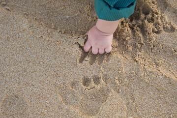 The baby is playing with the sand, making footprints in it. The baby's hand and foot are visible as they play on the beach.