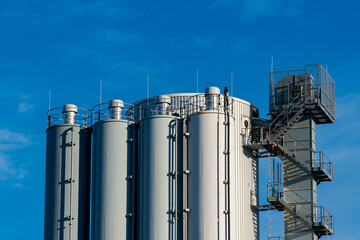 The view shows several tall industrial storage silos reaching towards a clear blue sky. A metal staircase leads to an observation platform situated on the side of one silo, indicating active use.