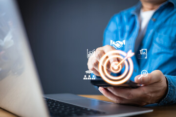 A businessman in a blue shirt interact with a smartphone, displaying holographic business icons, including a glowing target symbolizing goal achievement, analytics, partnerships, and financial growth.