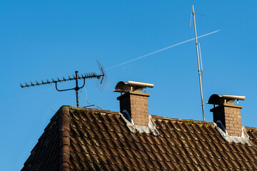 Two chimneys and an antenna stand on a sloped roof against a vibrant blue sky. A faint contrail...