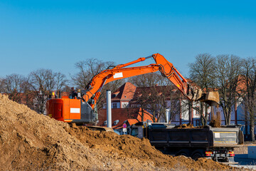 An excavator is loading soil from a large mound into a truck at a construction site. The clear blue sky and trees in the background enhance the daylight visibility of the machinery.