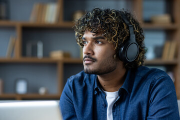 Young adult wearing headphones looks thoughtfully into the distance, wearing a blue denim shirt in front of a bookshelf, inside.