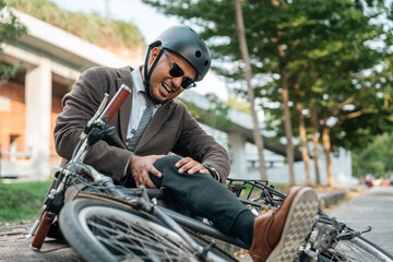 Obraz premium Asian Businessman falling off bicycle in city. Accident Injury leg pain. Broken leg Cyclist falls off the bike. Accident on the road.