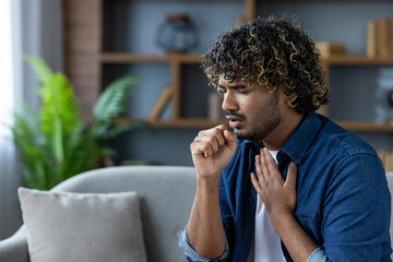 Man with curly hair coughing while sitting on a sofa in an interior setting, possibly experiencing...