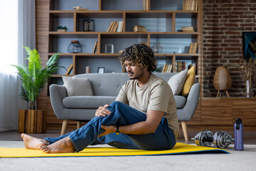 Young indian man sitting on a yoga mat at home, holding his injured knee, experiencing pain after a...