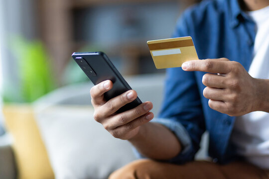 Man is holding a gold credit card and smartphone while sitting indoors. He is about to make an online payment using his mobile device. - Powered by Adobe