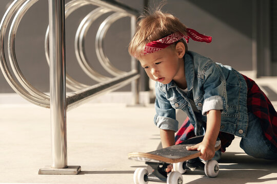 Delightful toddler boy wearing a denim outfit and red bandana plays with a skateboard in an urban setting